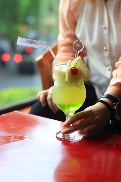 Eine Person sitzt an einem Tisch mit einem Glas grüner Flüssigkeit und einem Strohhalm, auf dem „Gurke“ steht.