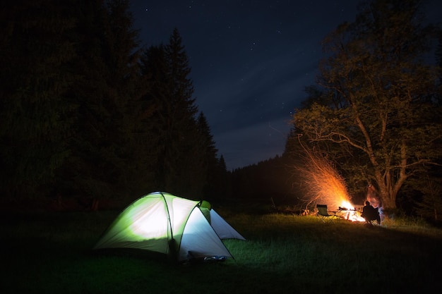 Eine Person sitzt bei einem Lagerfeuer, während sie nachts im Wald zeltet