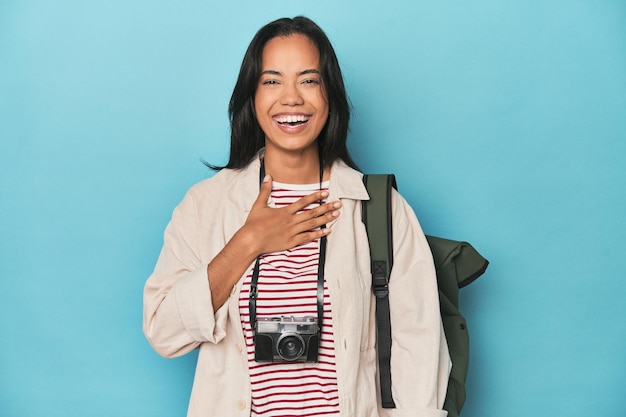Foto eine philippinische frau mit kamera und blauem rucksack lacht laut und hält ihre hand auf der brust