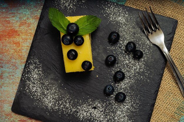 Eine Portion hausgemachter Lemon Curd Cake in Form eines Barrens oder Ziegels, dekoriert mit einigen Minzblättern und Blaubeeren auf einer schwarzen Platte. Ansicht von oben. Platz kopieren.