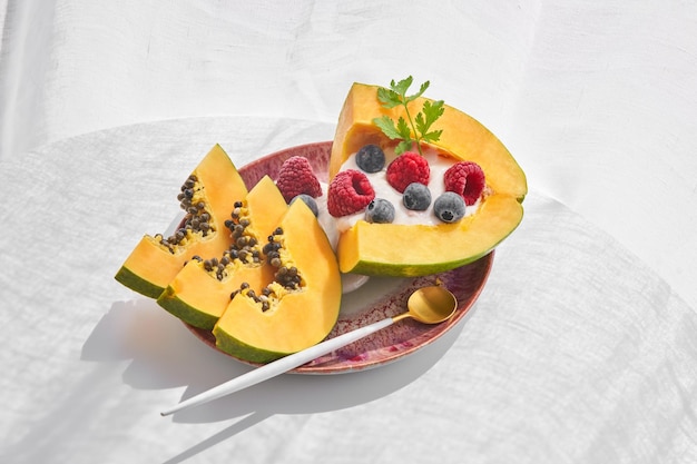 Eine Portion süße Papaya-Früchte, serviert auf einem rosa Keramikteller mit Löffel und dekoriert mit Joghurt und Beeren auf weißem Hintergrund