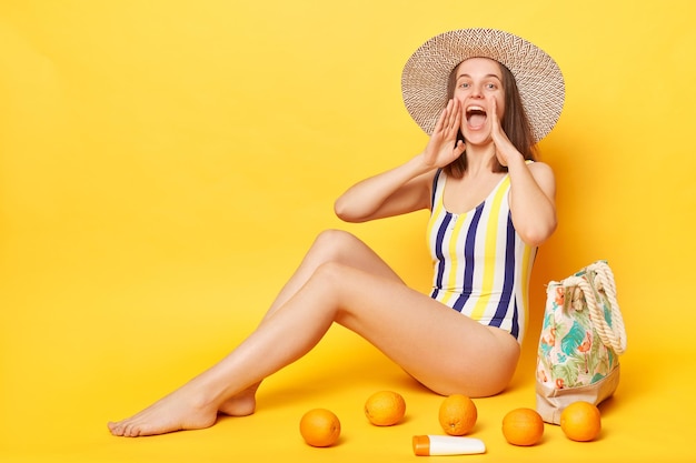 Eine positive Frau im einteiligen Badeanzug posiert isoliert vor gelbem Hintergrund und hält die Hände vor den Mund, schreit mit glücklichem Gesichtsausdruck und macht Ansagen