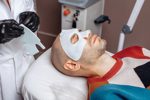 Eine professionelle Kosmetikerin stellt eine feuchtigkeitsspendende Gesichtsmaske zur Hautpflege mit zwei Teilen für einen jungen Mann her