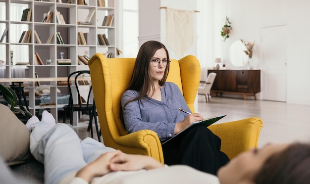 Eine Psychologin mit Brille sitzt in einem Sessel und berät einen Patienten auf der Couch. Professionelle psychologische Hilfe