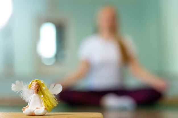 Eine Puppe in Form eines Engels auf unscharfem Hintergrund mit gesunder Frau, die Yoga im Innenbereich praktiziert