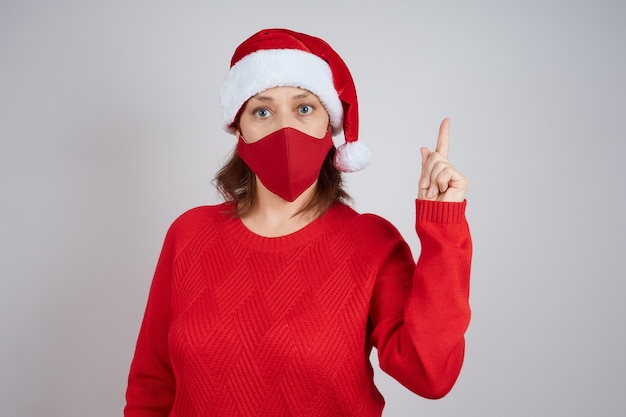 Eine reife Frau in einer roten Schutzmaske in einer Weihnachtsmütze auf grauem Hintergrund zeigt eine Geste der Aufmerksamkeit. Weihnachten, Neujahr in Quarantäne.