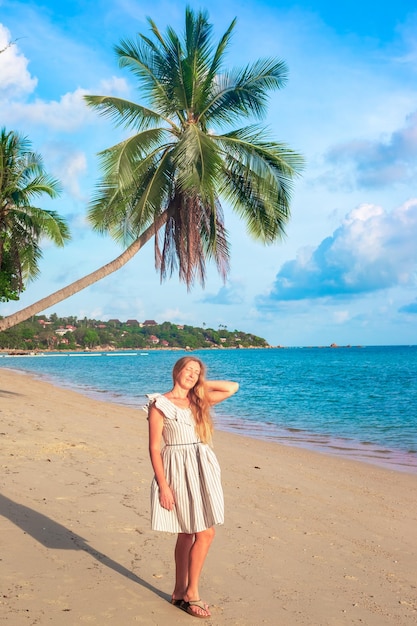 Eine reife Frau mit langen blonden Haaren in einem Kleid am Strand neben einer Palme genießt die Sonne