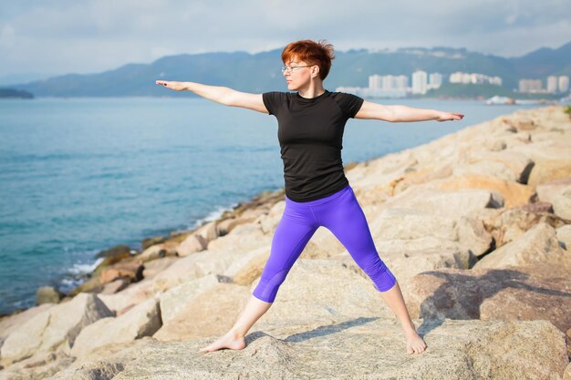 Eine reife Frau übt Yoga auf Felsen am Strand.