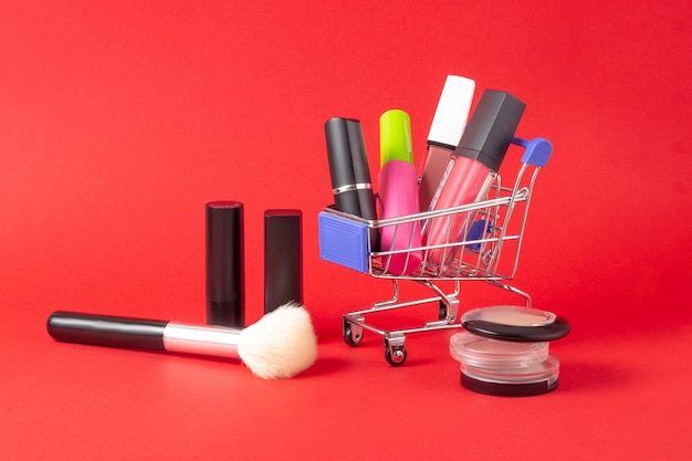 Eine Reihe von Lippenstiftkosmetik und Lipgloss, Puder, Lidschatten und ein Einkaufswagen auf leuchtend rotem Hintergrund. Das Konzept des Kaufs von Kosmetika, Online-Shopping, Urlaub