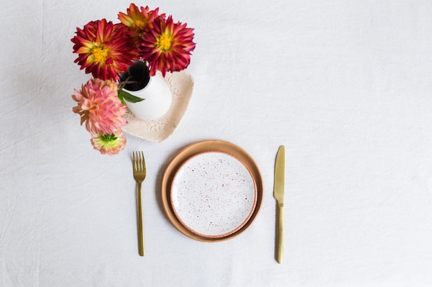 Eine Reihe von orangefarbenen Tonplatten, ein Messer und eine Gabel auf einer weißen Tischdecke aus Naturmaterial. Eine Vase mit Dahlienblüten steht in der Nähe. Draufsicht