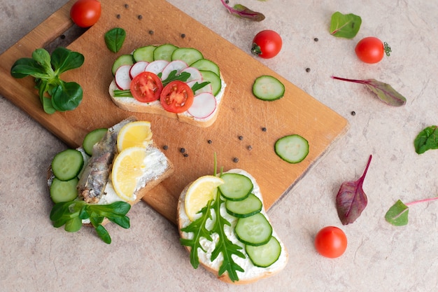 Eine Reihe von Sandwiches mit Sprotten, hausgemachter Quarkcreme mit Kräutern, mit Radieschen, Gurken und Tomaten auf einem hölzernen Hintergrund. Ansicht von oben. Platz kopieren.