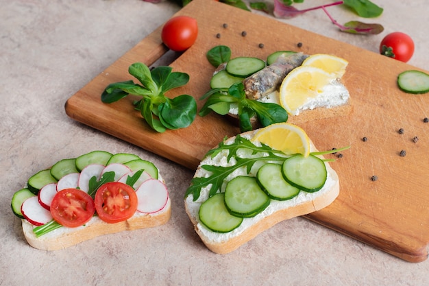 Eine Reihe von Sandwiches mit Sprotten, hausgemachter Quarkcreme mit Kräutern, mit Radieschen, Gurken und Tomaten auf einem hölzernen Hintergrund. Ansicht von oben. Platz kopieren.