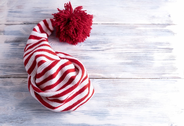 Eine rot-weiß gestreifte Kappe mit einem langen Schwanz und einem großen roten Pompon befindet sich auf einem weißen Holztisch, flach gelegen, Draufsicht.