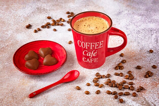 Eine rote Tasse Kaffee mit einem Löffel Kaffeebohnen und Pralinen auf einem roten Teller auf hellem Hintergrund