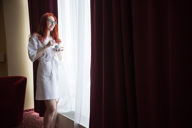 Eine rothaarige Frau, die im Hotelzimmer am Fenster steht