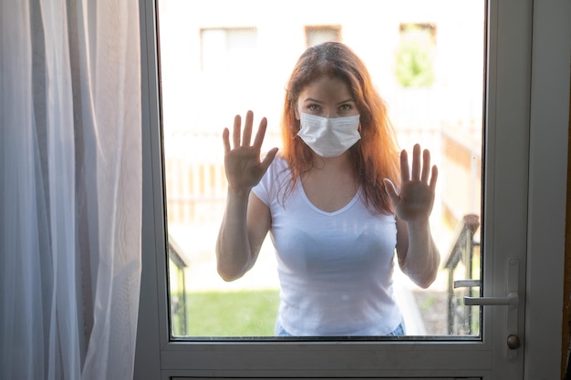 Eine rothaarige Frau mit medizinischer Maske lehnte an der vorderen Glastür. Ein Mädchen steht während der Quarantäne-Coronavirus-Epidemie vor dem Fenster. Soziale Distanz