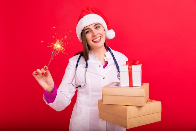 Eine Ärztin in einem Weihnachtsmann-Hut hält Schachteln mit Geschenken und eine Wunderkerze auf rotem Hintergrund Herzlichen Glückwunsch des Arztes an die PatientenxAxAxA