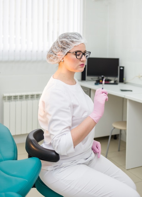 Eine Ärztin in einem weißen medizinischen Anzug, die Kappe sitzt seitlich und hält eine Spritze zur Injektion im Büro