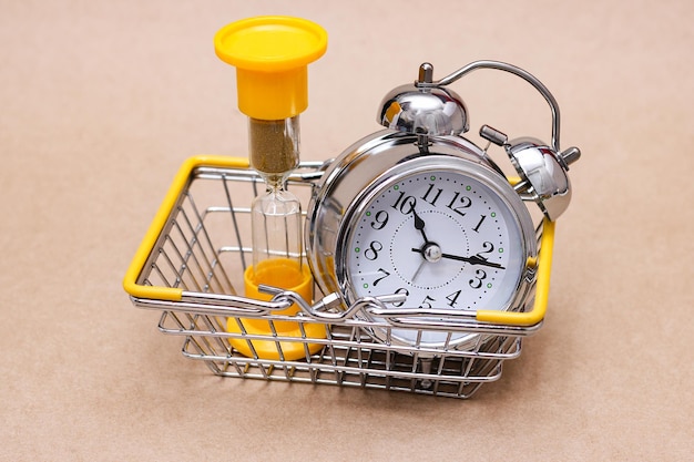Eine Sanduhr und ein Wecker aus Metall im Einkaufskorb. Konzept der Zeit.