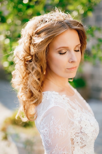 Eine sanfte blondhaarige Braut in einer Hochzeitstiara steht mit geschlossenen Augen im Sonnenlicht