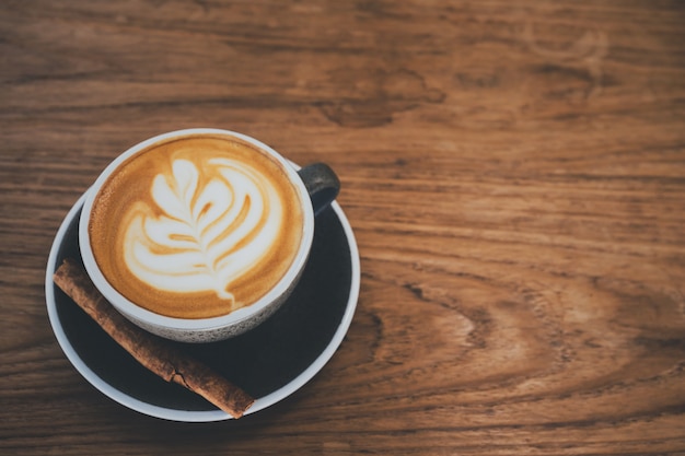 Eine Schale Cappuccinokaffee mit Lattekünsten auf Holztisch im Kaffeestubehintergrund mit Weinlesefilter.