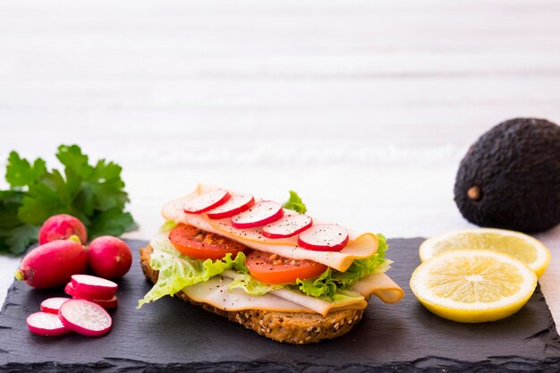 Eine Scheibe Vollkornbrot mit Kernen, gebratenem Truthahn und frischem Gemüse der Saison, auf einem schwarzen Steinbrett