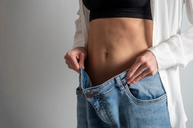 Eine schlanke Frau mit nacktem Bauch zeigt, wie sie abgenommen hat und hält ihre Jeans mit der Hand. Das Konzept der Ernährung, Gewichtsverlust, gesunde Lebensweise