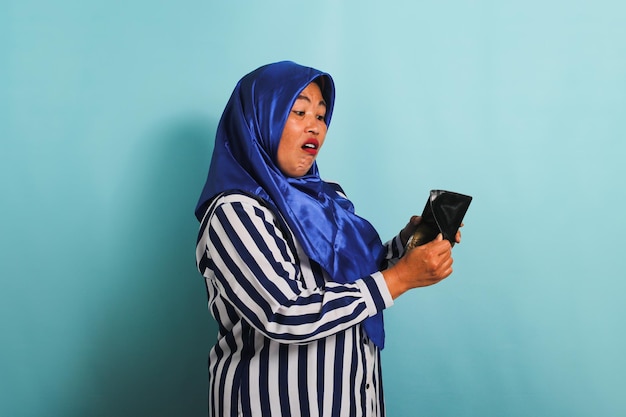 Eine schockierte asiatische Frau mittleren Alters im Hijab schaut in ihre leere Brieftasche, isoliert auf blauem Hintergrund.
