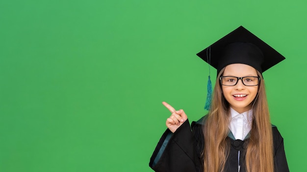 Eine schöne Absolventin in einer Kappe und einem zeremoniellen Gewand zeigt mit ihrer Hand auf einen isolierten grünen Hintergrund. Erwerb eines Abschlusszeugnisses in der Grundschule.