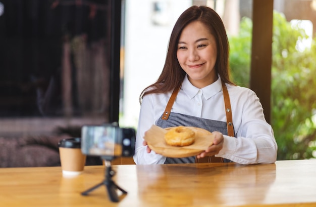 Eine schöne asiatische Foodbloggerin oder Vloggerin, die ein Stück Donut zeigt, während sie ein Video mit der Kamera aufnimmt