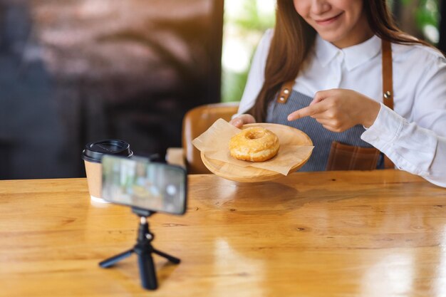 Eine schöne asiatische Foodbloggerin oder Vloggerin, die ein Stück Donut zeigt, während sie ein Video mit der Kamera aufnimmt