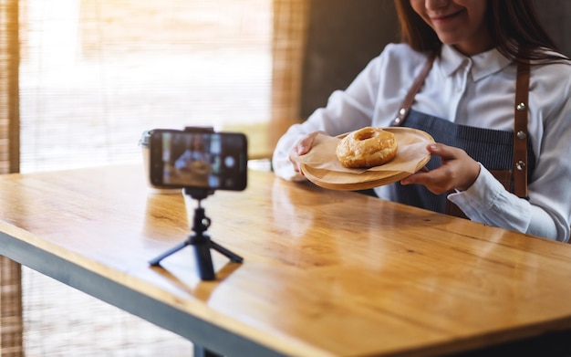 Eine schöne asiatische Foodbloggerin oder Vloggerin, die ein Stück Donut zeigt, während sie ein Video mit der Kamera aufnimmt