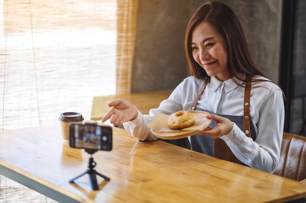 Eine schöne asiatische Foodbloggerin oder Vloggerin, die ein Stück Donut zeigt, während sie ein Video mit der Kamera aufnimmt