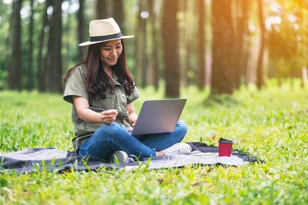 Eine schöne asiatische Frau, die auf der Laptoptastatur arbeitet und tippt, während sie im Park sitzt