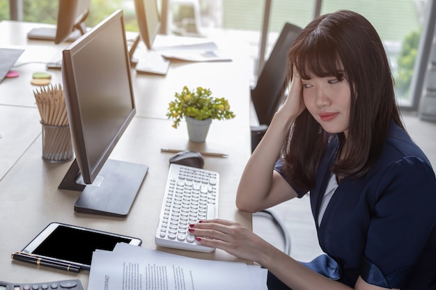 Eine schöne asiatische Frau, die einen dunkelblauen Anzug trägt, der an einem Schreibtisch in einem modernen Büro sitzt und Stress arbeitet