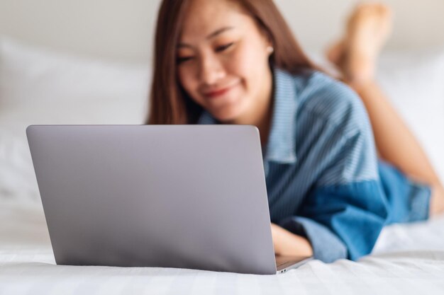 Eine schöne asiatische Frau, die einen Laptop verwendet und daran arbeitet, während sie sich zu Hause auf einem weißen, gemütlichen Bett hinlegt