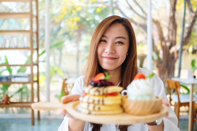 Eine schöne asiatische Frau, die einen Teller Pfannkuchen mit Eis und Schlagsahne im Café hält und zeigt