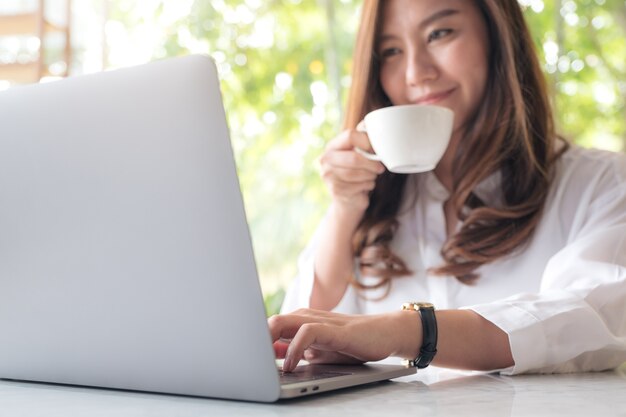 eine schöne asiatische Frau, die Laptop beim Trinken des Kaffees im Café verwendet