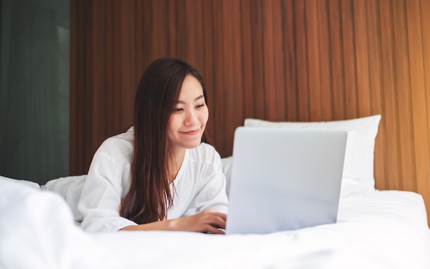 Eine schöne asiatische Frau, die Laptop-Computer benutzt und am Laptop arbeitet, während sie zu Hause auf einem weißen gemütlichen Bett liegt