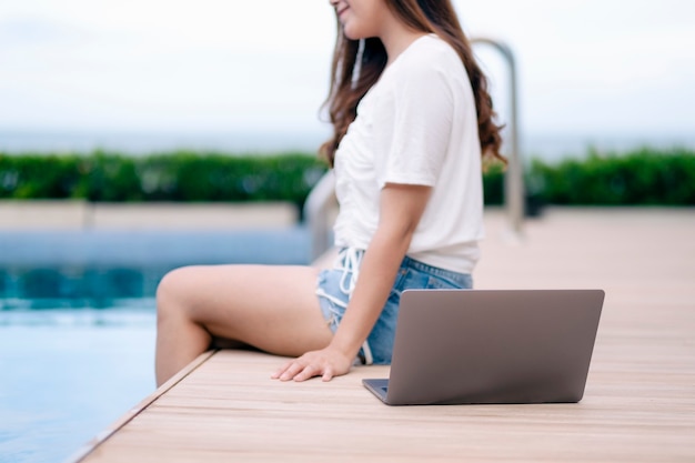 Eine schöne asiatische frau, die mit laptop-computer am schwimmbad sitzt