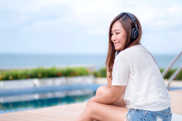 Eine schöne asiatische frau, die musik mit kopfhörer hört, während sie am schwimmbad sitzt