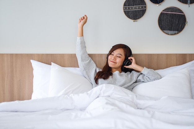 Eine schöne asiatische Frau hört gerne Musik mit Kopfhörer, während sie zu Hause auf einem weißen gemütlichen Bett liegt