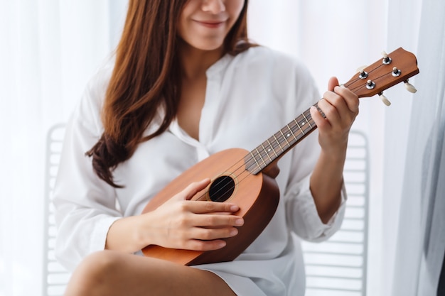 Eine schöne asiatische Frau sitzt und spielt Ukulele im Schlafzimmer