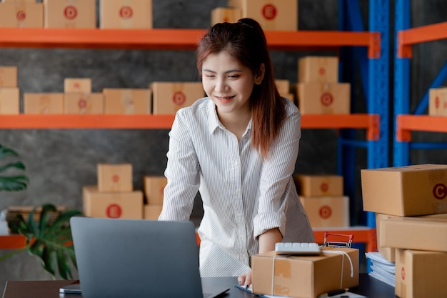 Eine schöne asiatische Geschäftsinhaberin eröffnet einen Online-Shop, in dem sie Bestellungen von Kunden prüft, die Waren über ein Kurierunternehmenskonzept einer Frau versenden, die ein Online-Geschäft eröffnet