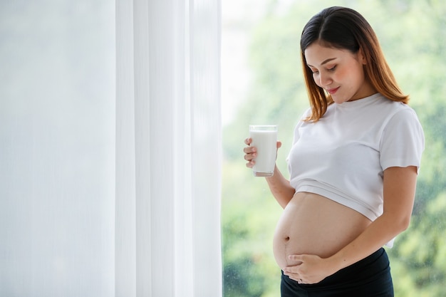 Eine schöne asiatische schwangere Frau, die ein weißes T-Shirt und eine schwarze Hose trägt, die am Fenster steht und ein Glas Milch hält, während sie ihren Bauch mit Liebe betrachtet. Konzept der Familie Mutter.