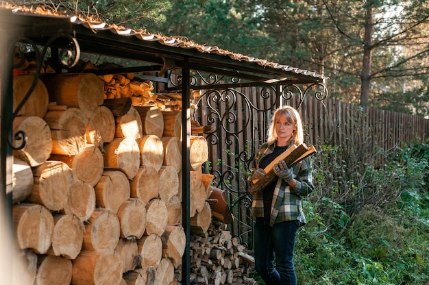 Eine schöne blonde Frau in einem karierten Hemd sammelt Holz von einem Holzofen im ländlichen Alltag