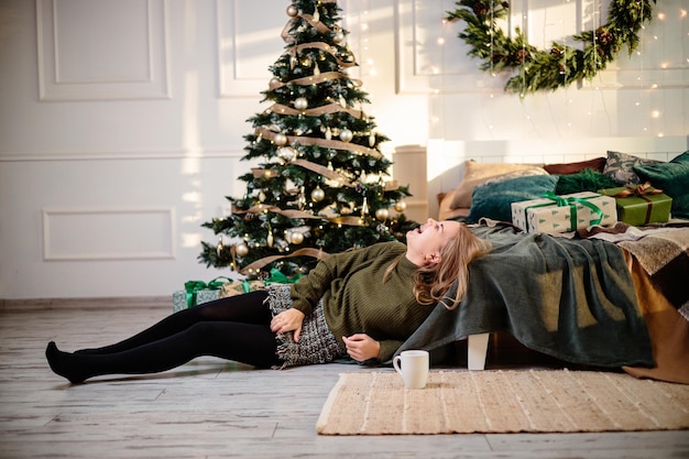 Eine schöne blonde Frau sitzt neben einem Weihnachtsbaum in einer Neujahrsatmosphäre und freut sich Die Frau befindet sich in einem hellen, komfortablen Schlafzimmer, das wunderschön für Weihnachten dekoriert ist