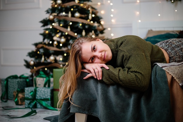 Eine schöne blonde Frau sitzt neben einem Weihnachtsbaum in einer Neujahrsatmosphäre und freut sich Die Frau befindet sich in einem hellen, komfortablen Schlafzimmer, das wunderschön für Weihnachten dekoriert ist