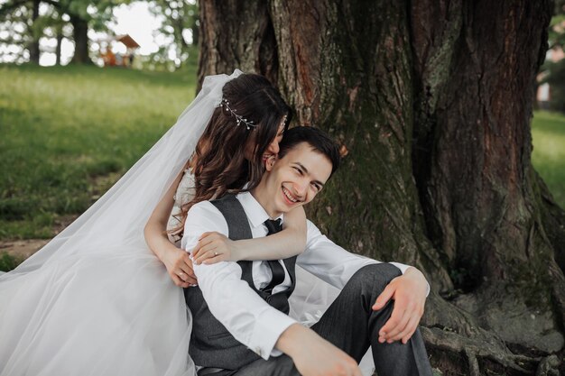 Eine schöne Braut mit langen lockigen Haaren in einem schicken Kleid umarmt den Bräutigam, lächelt und schaut in die Linse unter einem großen Baum. Porträt der Braut und des Bräutigen Frühlingshochzeit Natürliches Make-up