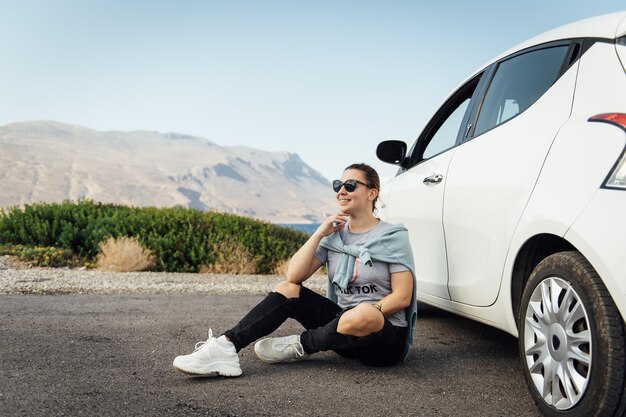 Eine schöne Brünette mit Sonnenbrille sitzt neben einem weißen Auto vor dem Hintergrund des Mittelmeers und der Berge